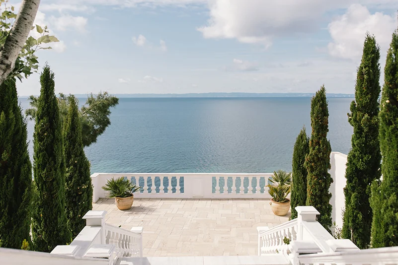 A peaceful view of the Aegean sea from a white stone terrace, framed by tall cypress trees in Halkidiki.