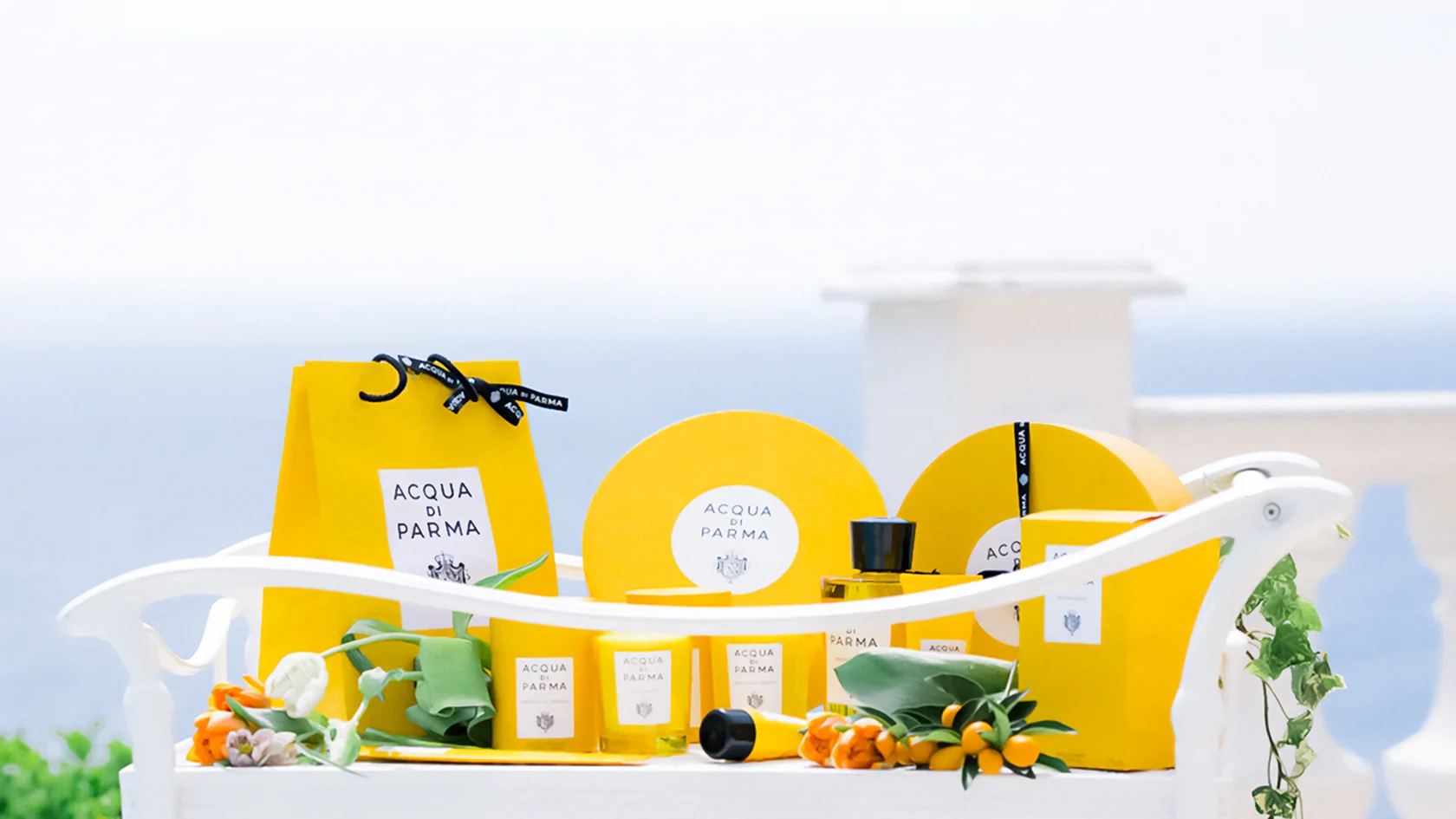 Bright yellow Acqua di Parma gift packaging and candles arranged on a white outdoor bench with flowers and greenery.