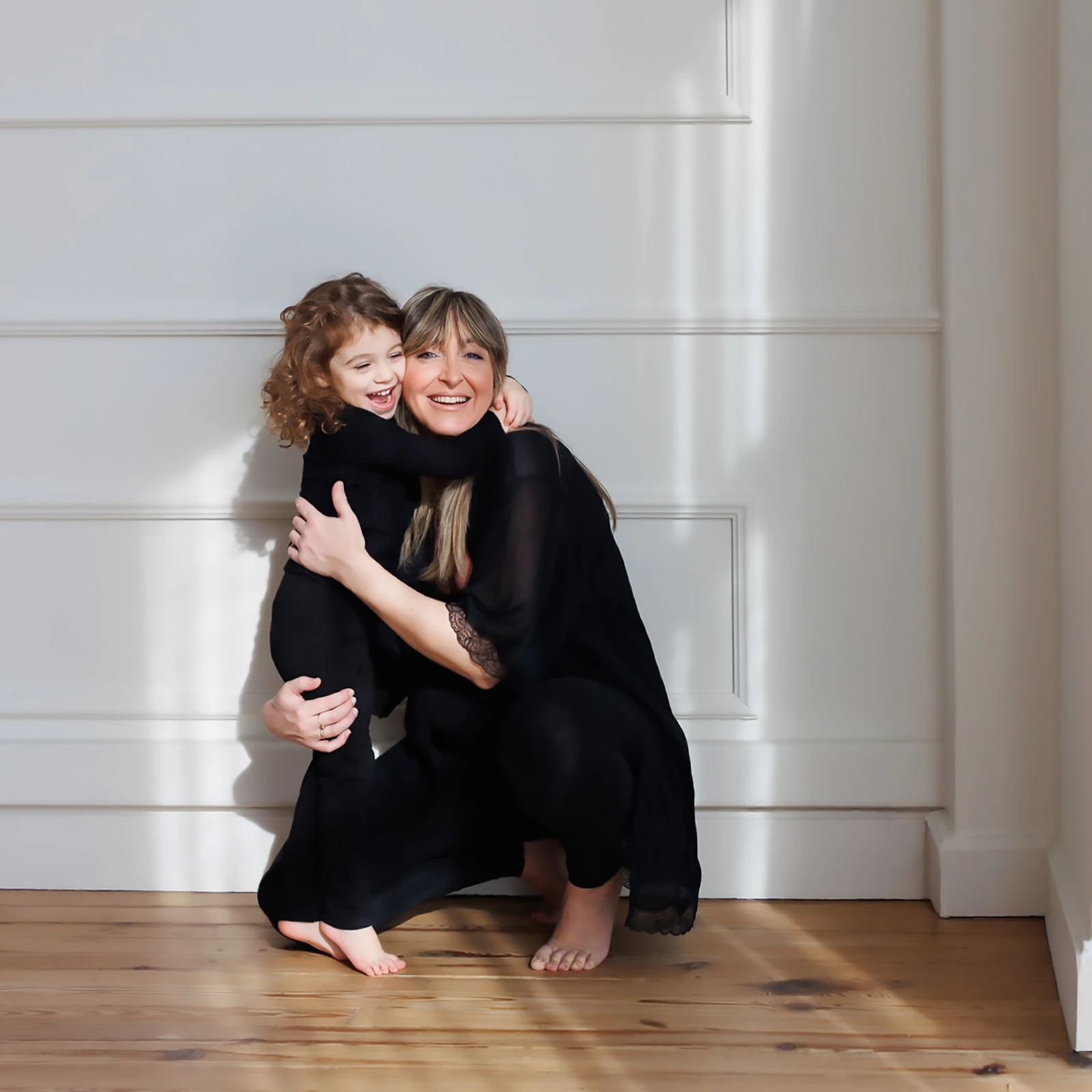 Smiling woman crouching and hugging a laughing young child near a white wall on wooden floor.