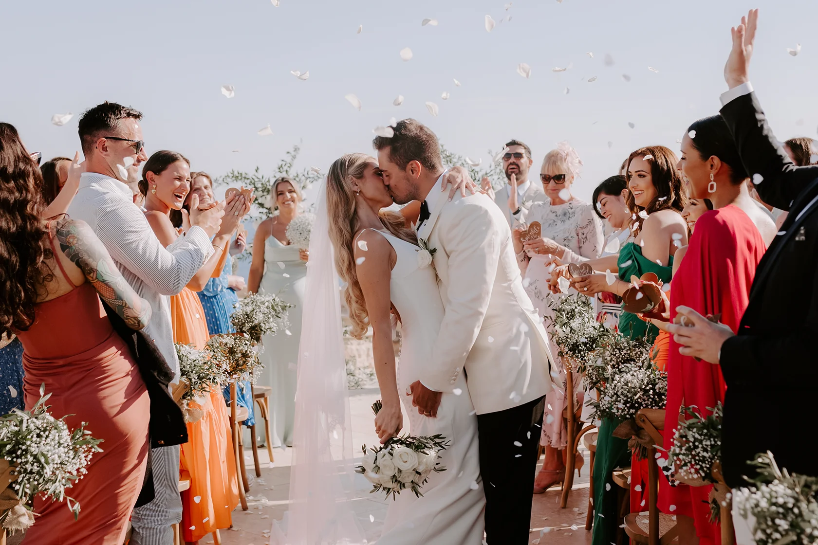 Bride and groom kissing at outdoor wedding ceremony aisle as guests clap and throw flower petals.