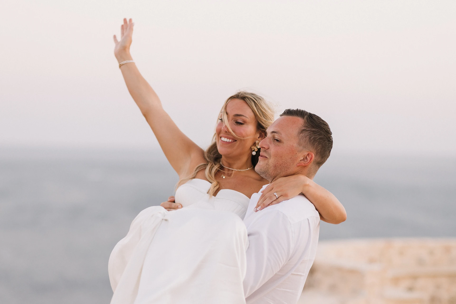 Man in white shirt carrying smiling woman in white dress with arm raised against a blurred outdoor background.