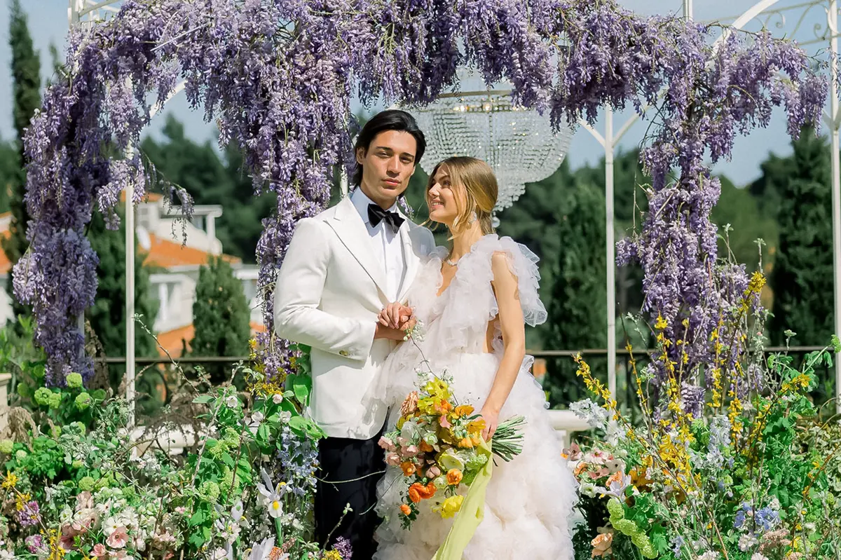 Bride and groom standing under a floral arch with purple flowers and greenery, bride holding a colorful bouquet.