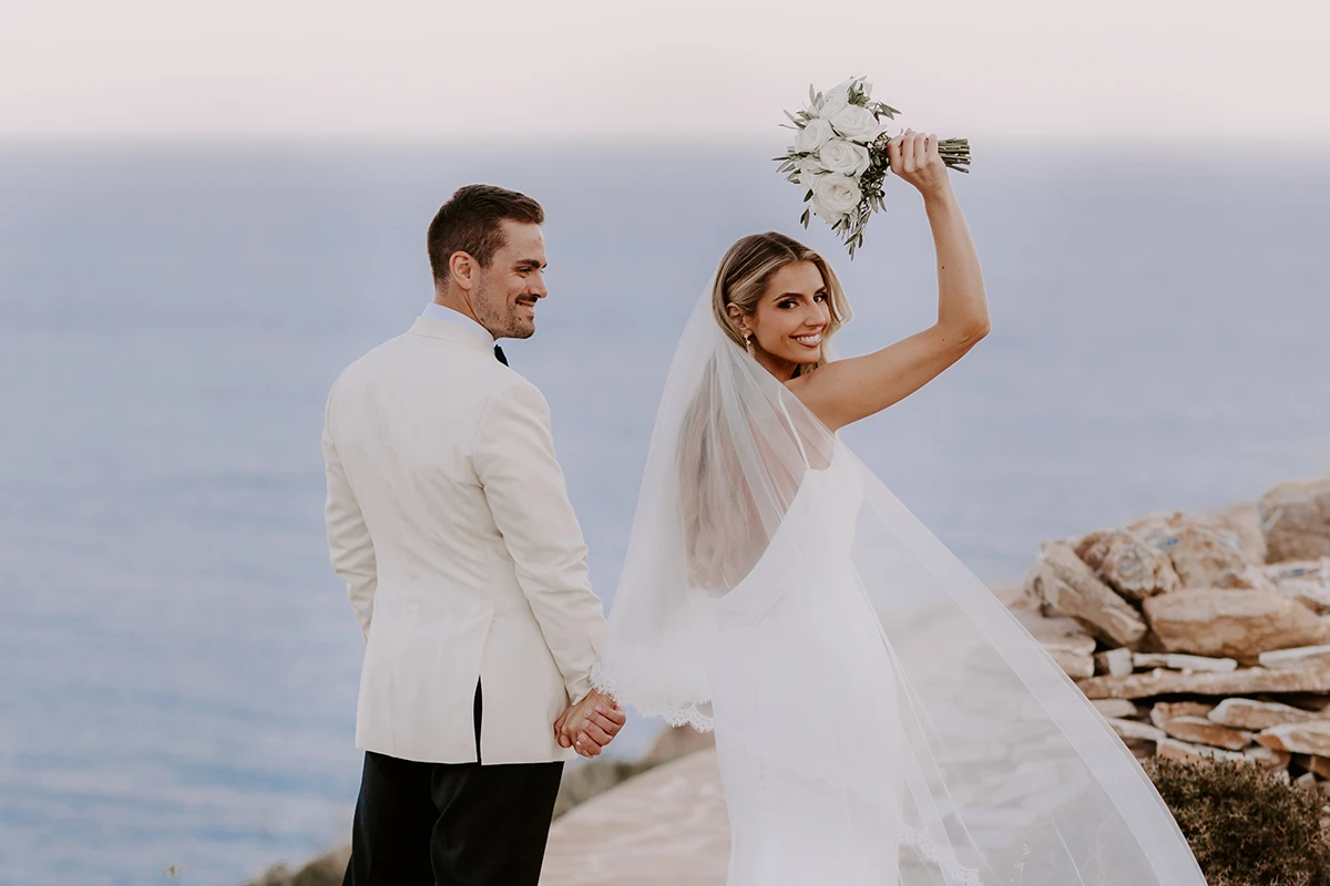 Smiling bride holding bouquet up and holding hands with groom by rocky seaside.