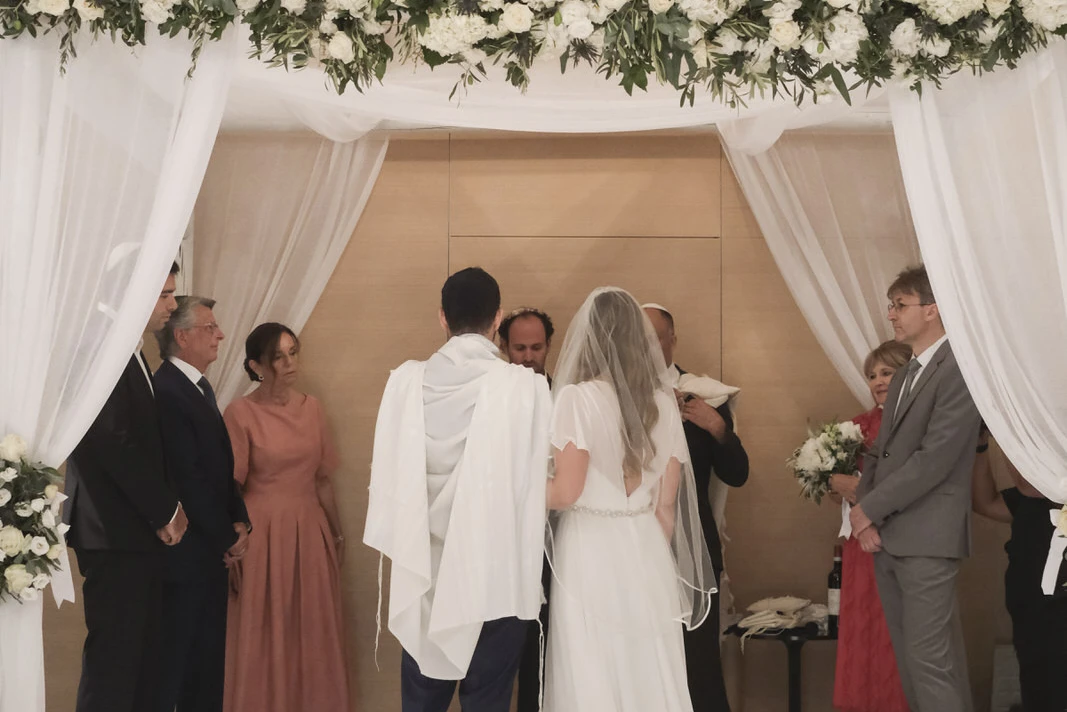Bride in veil and groom stand under floral wedding arch, facing officiants during ceremony.