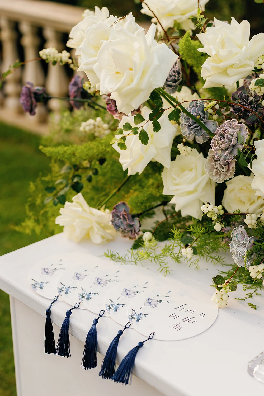 White and purple floral arrangement on a table with round fans featuring navy tassels and script text.