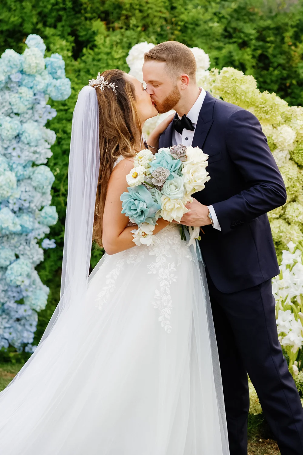 Bride and groom kissing outdoors surrounded by white and blue floral arrangements, bride holding a bouquet.