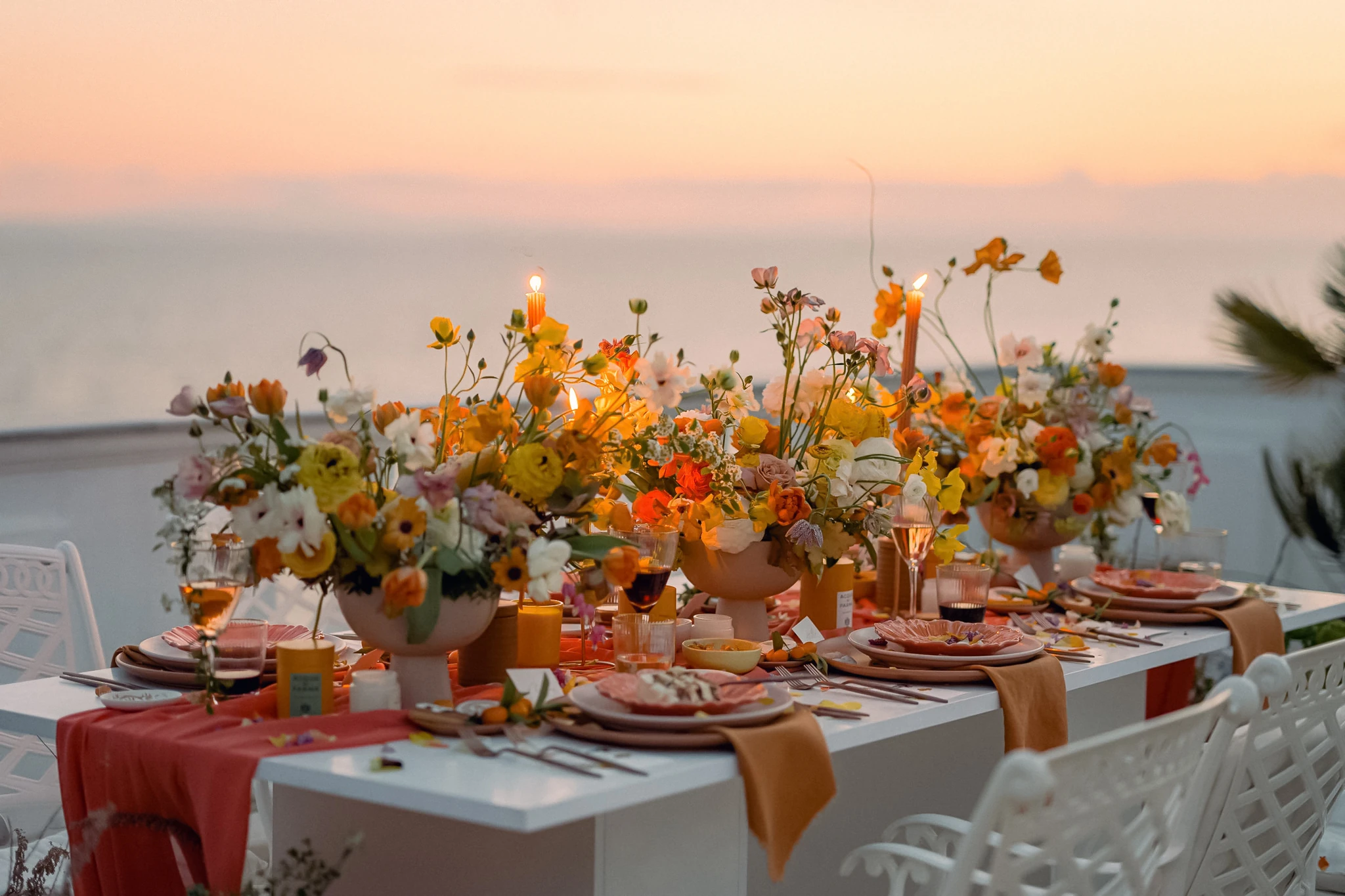 Elegantly set outdoor dining table with vibrant floral arrangements and candles at sunset.