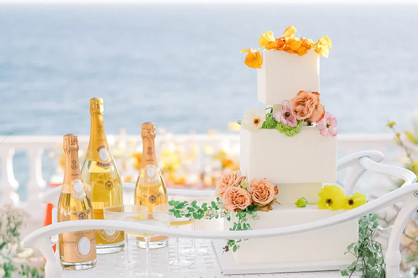 Three-tier white wedding cake decorated with orange and pink flowers on a white table near ocean with champagne bottles.