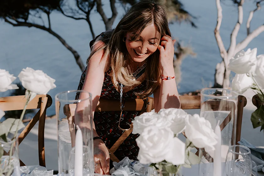 Smiling woman arranging white roses on an outdoor table by the water.