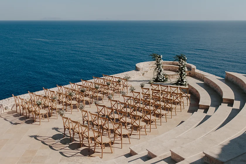 Outdoor wedding setup with wooden chairs facing floral arch overlooking the Aegean Sea.