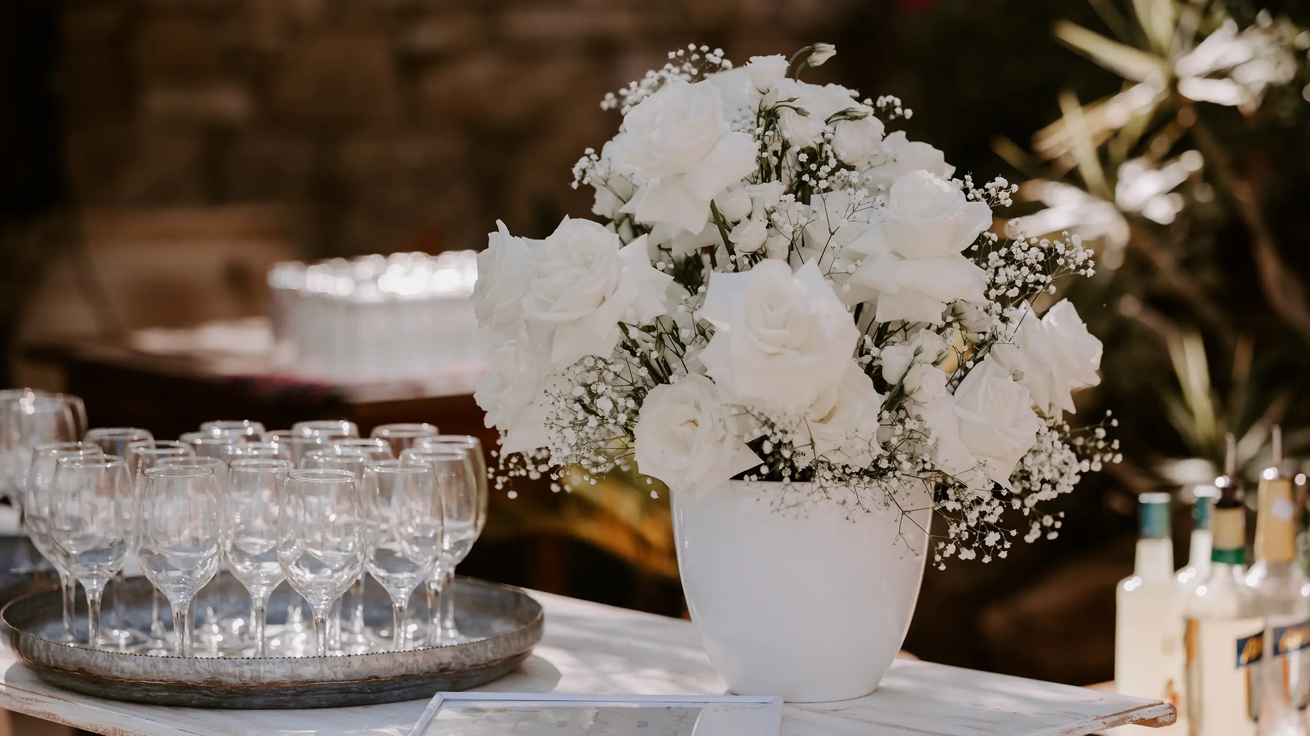 White rose bouquet in vase next to a tray of empty wine glasses on a table.