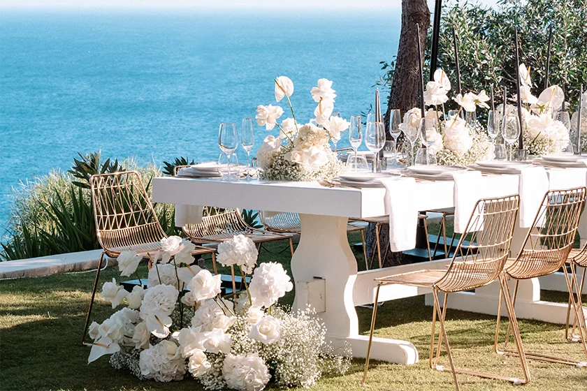 Elegant outdoor dining table with white flowers and glassware facing the Aegean Sea under clear sky.