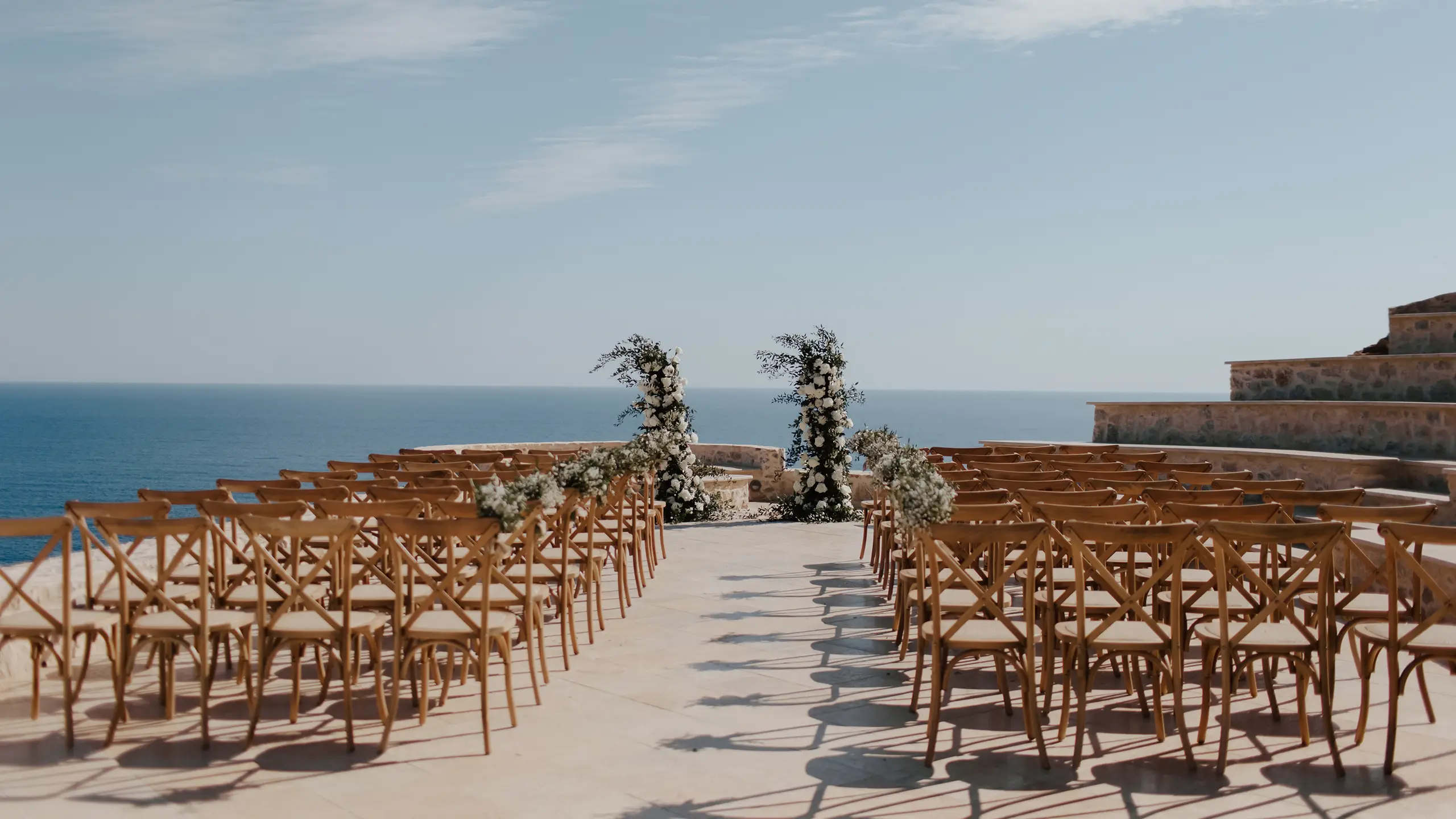 Outdoor wedding setup with wooden chairs and floral arches by the sea under a clear sky.