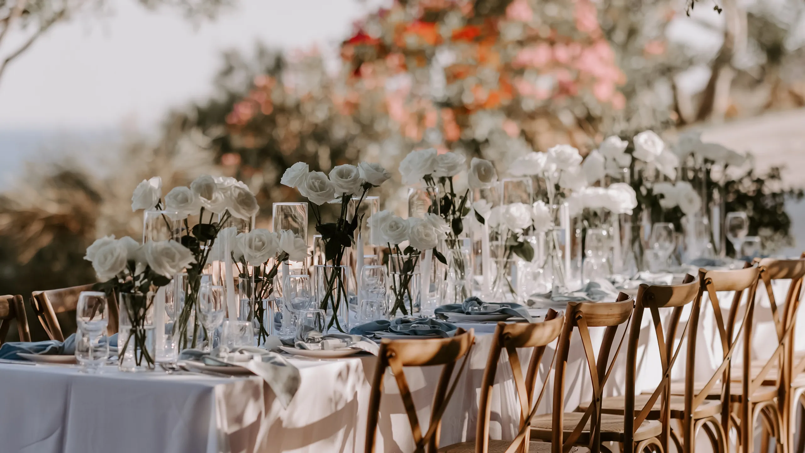 Outdoor dining table with white roses in vases, glassware, and wooden chairs set for an event.