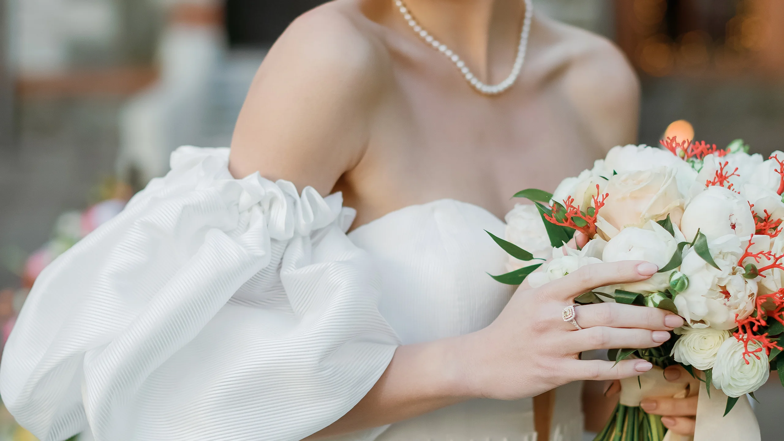 Bride in white dress with pearl necklace holding a bouquet of white and red flowers.