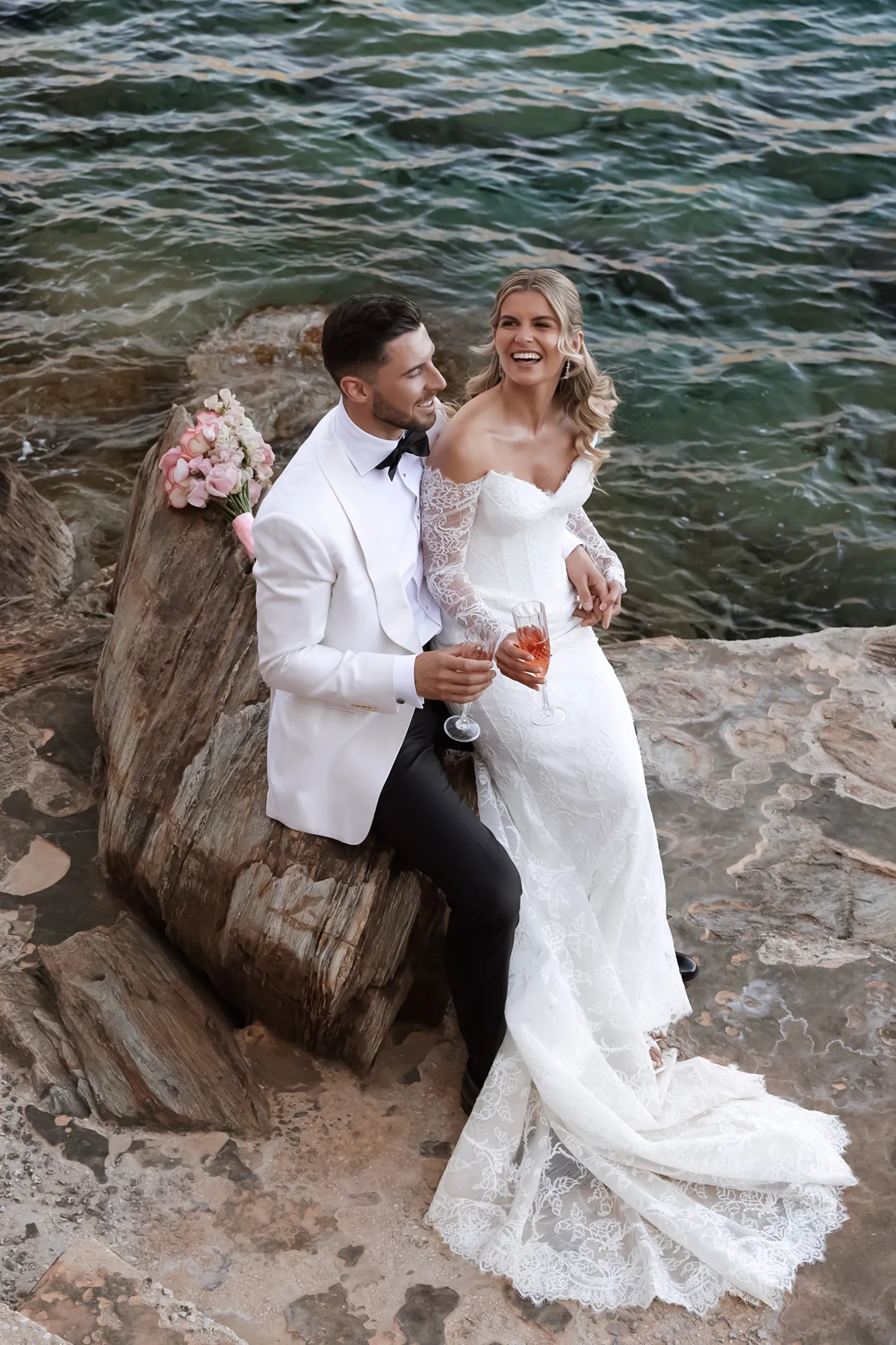 Bride in lace dress and groom in white jacket sitting on rocks by water holding glasses of rose wine.
