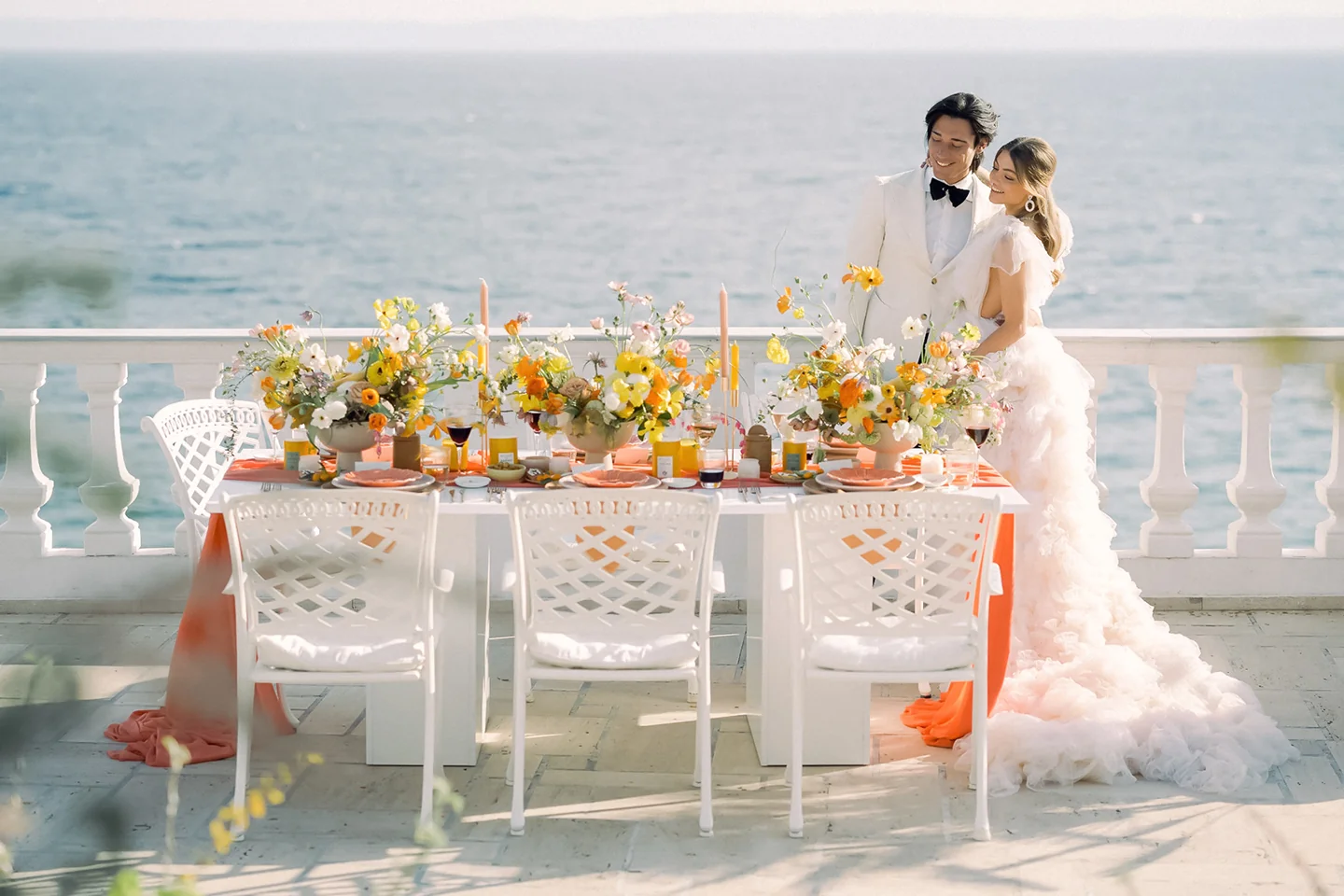 Bride and groom in elegant attire stand by a floral-decorated table with ocean view in the background.