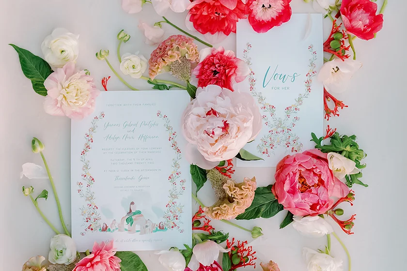 Two floral wedding invitation cards surrounded by pink, red, and white flowers on a white surface.