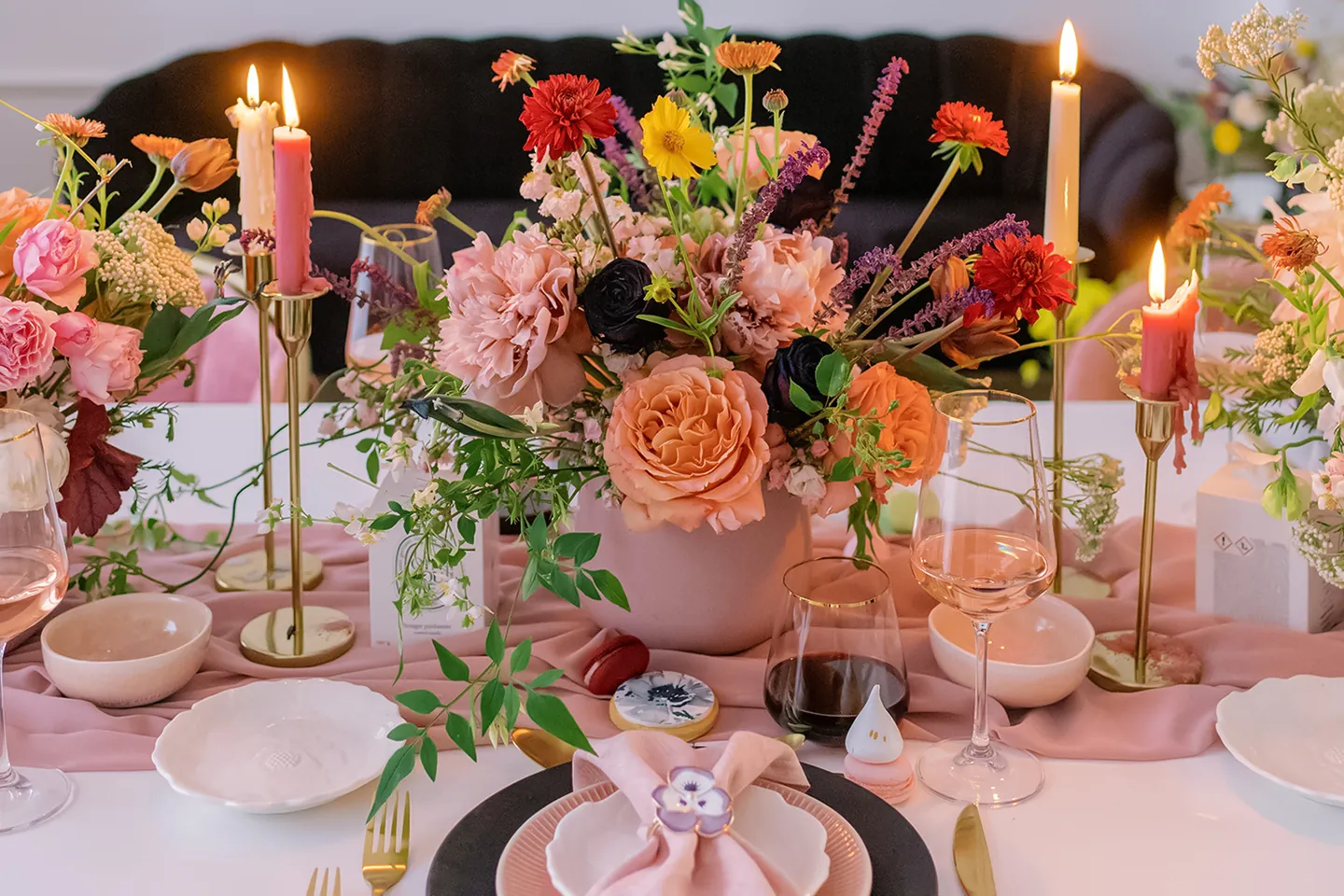 Elegant table set with floral centerpiece, pink napkin, candles, wine glasses, and gold cutlery on white table.