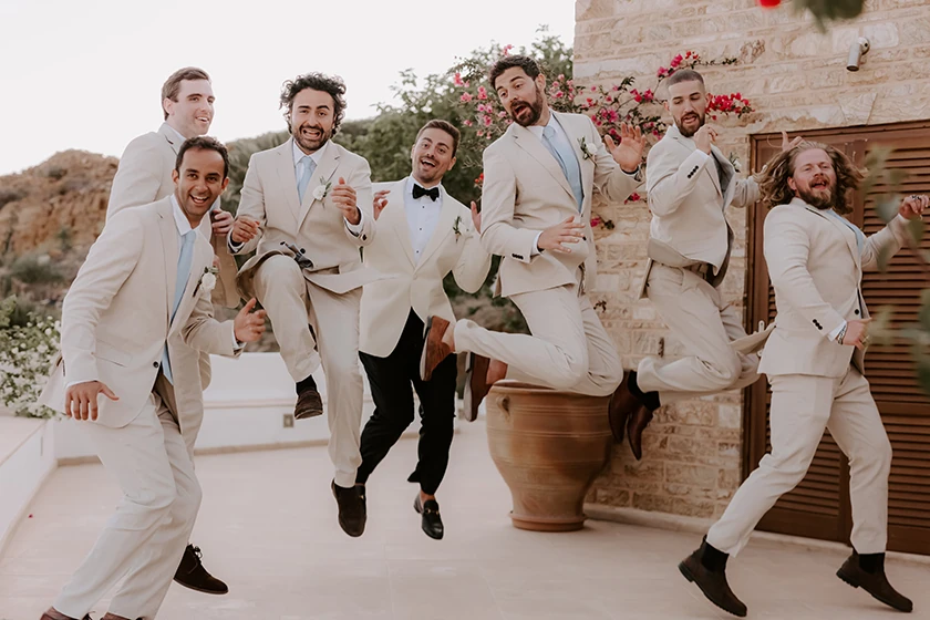 Seven men in light suits and one in a black tuxedo jumping joyfully outdoors near a stone wall with flowers.
