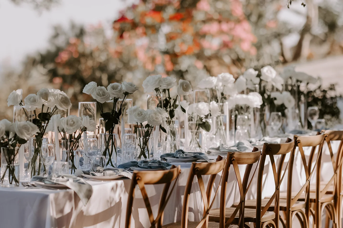Elegant outdoor table set with white roses in glass vases, glassware, and wooden chairs ready for event.