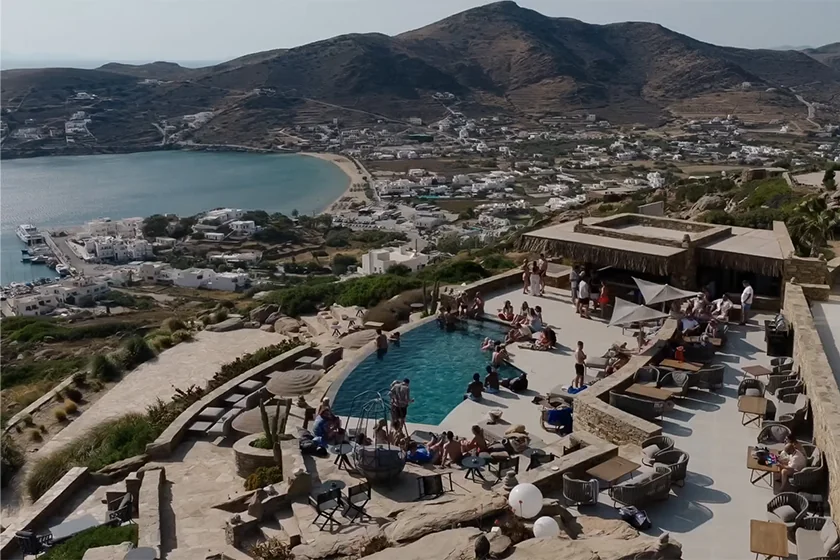 Outdoor pool area with people relaxing and swimming, overlooking a coastal town and hills in the distance.