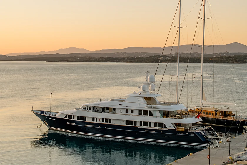 Large luxury yacht docked at a harbor during sunset with calm water and distant hills.