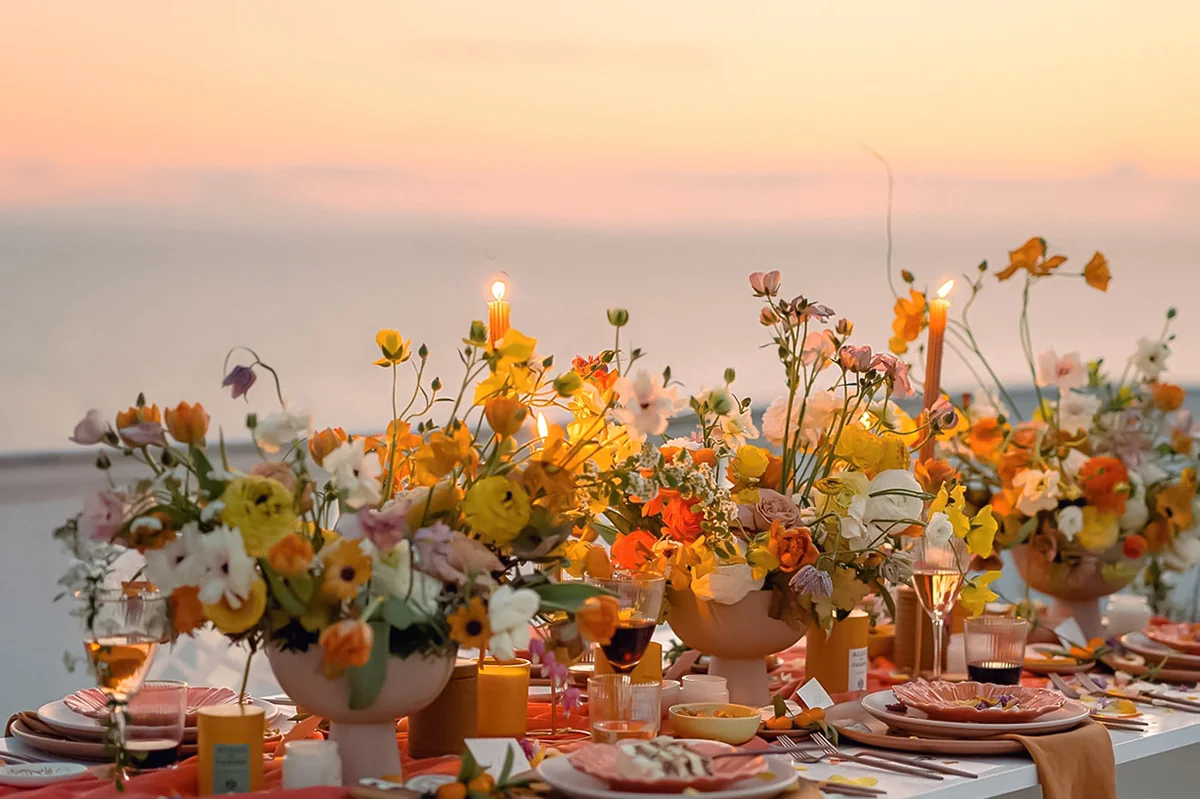 Elegant table setting with vibrant flower centerpiece, lit candles, and glassware at sunset.