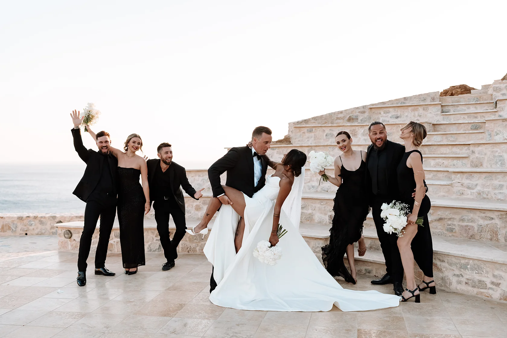 Groom dipping bride holding bouquet as friends cheer beside stone steps by the sea at sunset on Ios island.