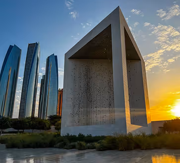 Modern monument with geometric cutouts at sunset, with tall glass skyscrapers in the background.