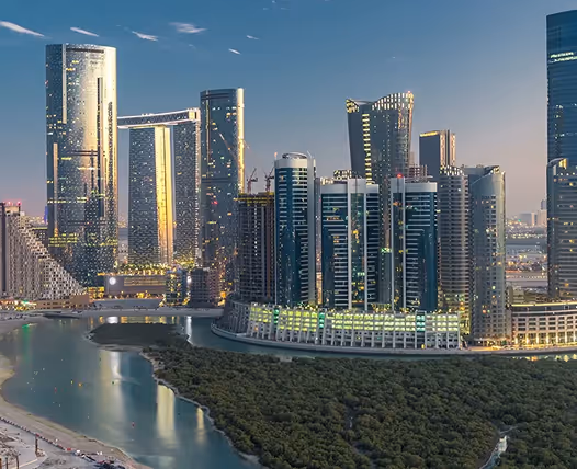 Twilight view of a modern city skyline with tall skyscrapers reflecting light and dense green trees along a waterfront.