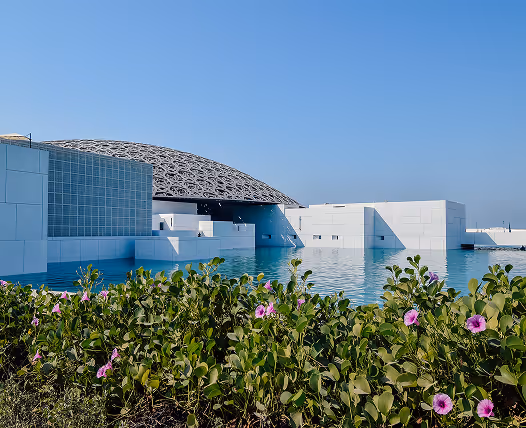 Modern white building with a large intricate dome roof, surrounded by a reflective water pool and flowering green shrubs under a clear blue sky.