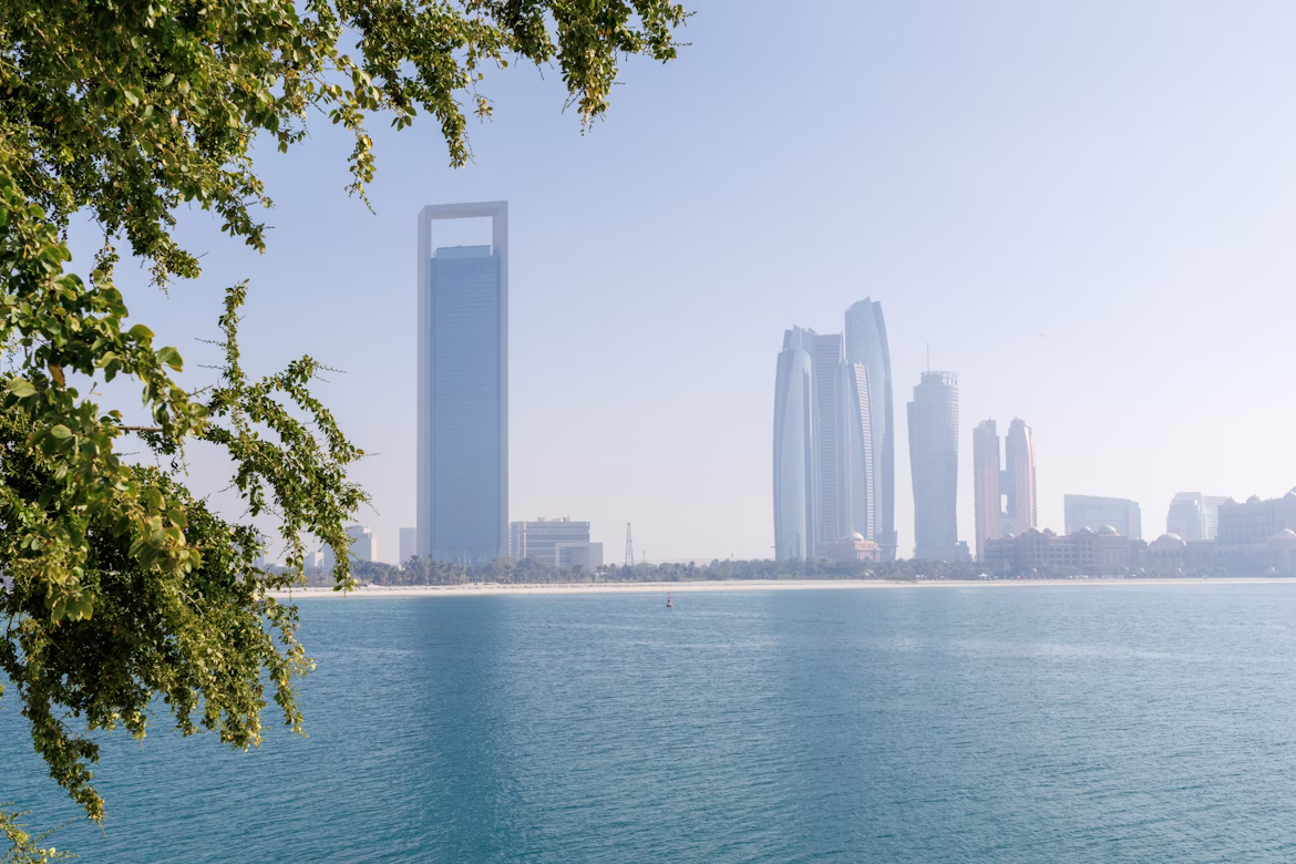 View of Abu Dhabi skyline with the Landmark Tower and Etihad Towers seen across calm blue water framed by green tree branches.