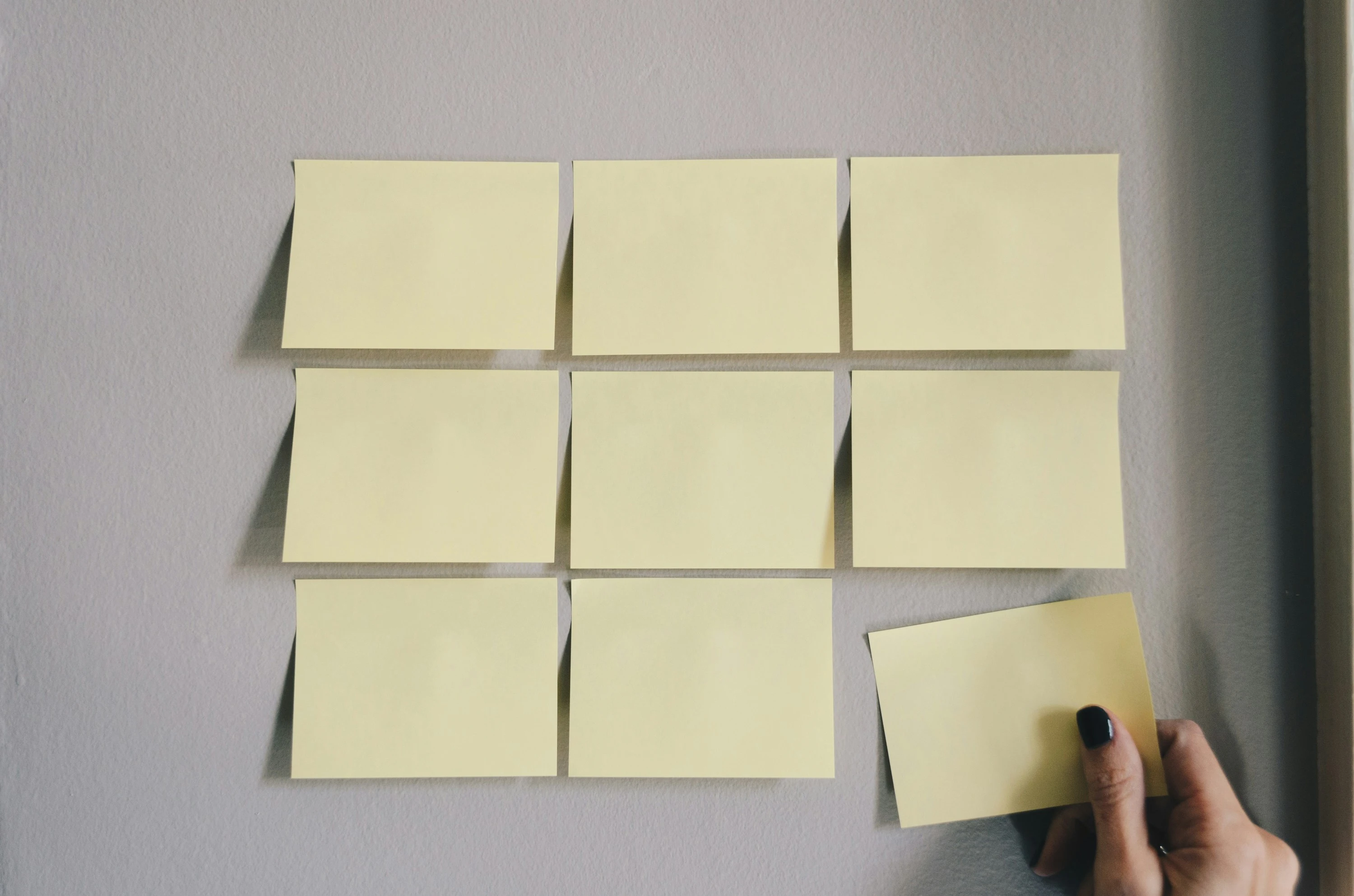 Hand placing a blank yellow sticky note on a wall with eight other blank sticky notes arranged in three rows.
