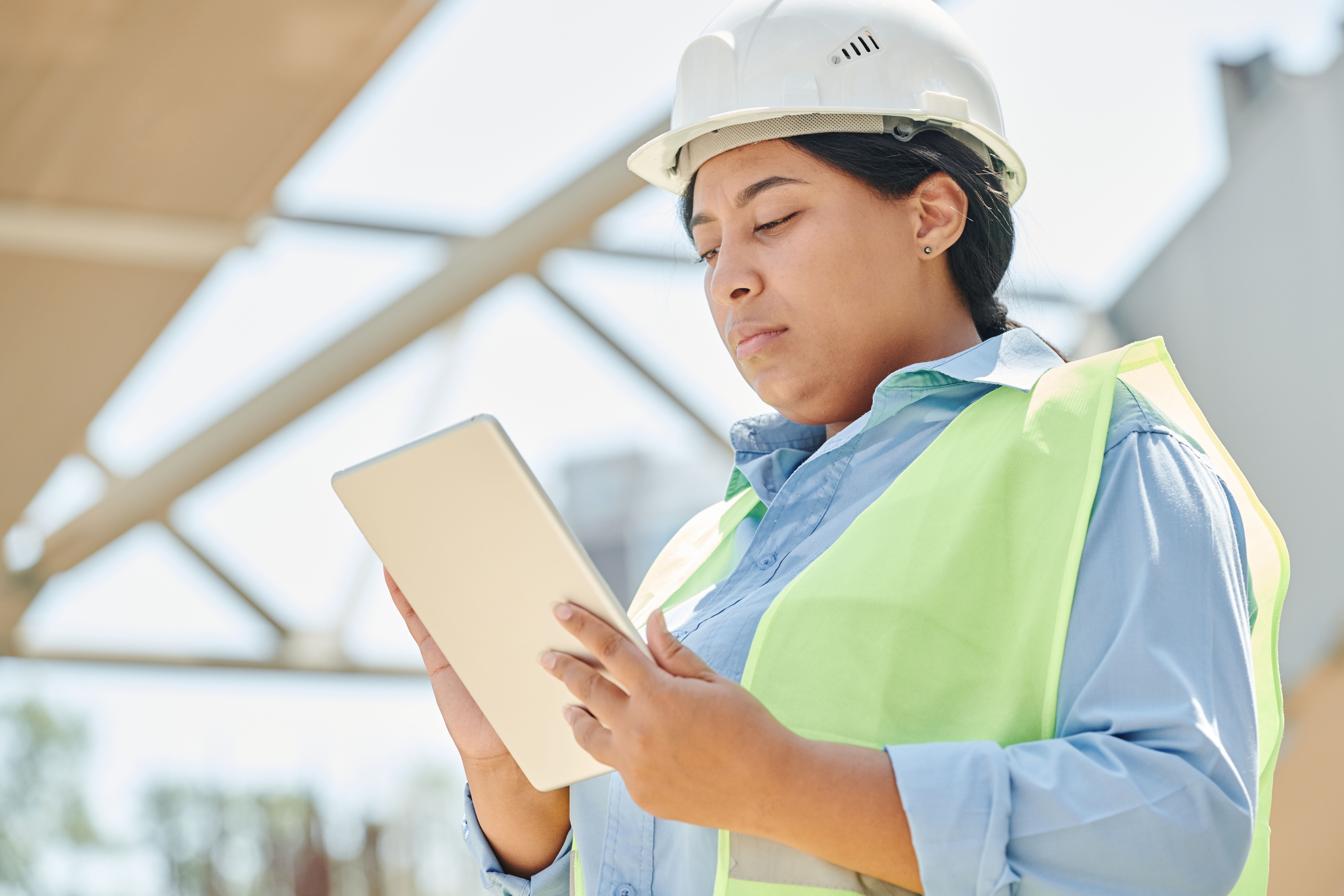 Construction worker wearing a white hard hat and yellow vest using a tablet at a worksite.