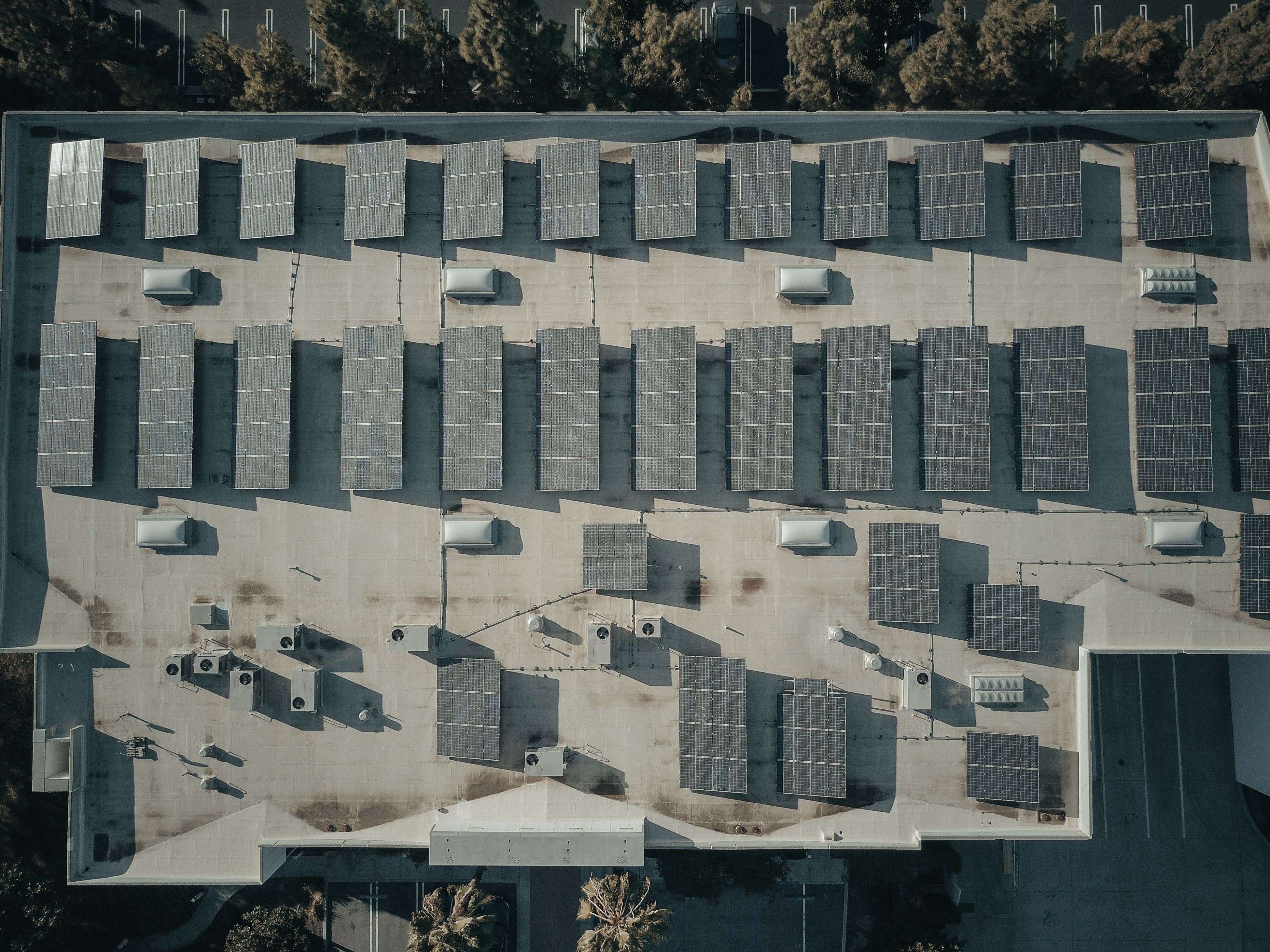 Aerial view of rectangular solar panels arranged on a large flat rooftop with shadows visible.