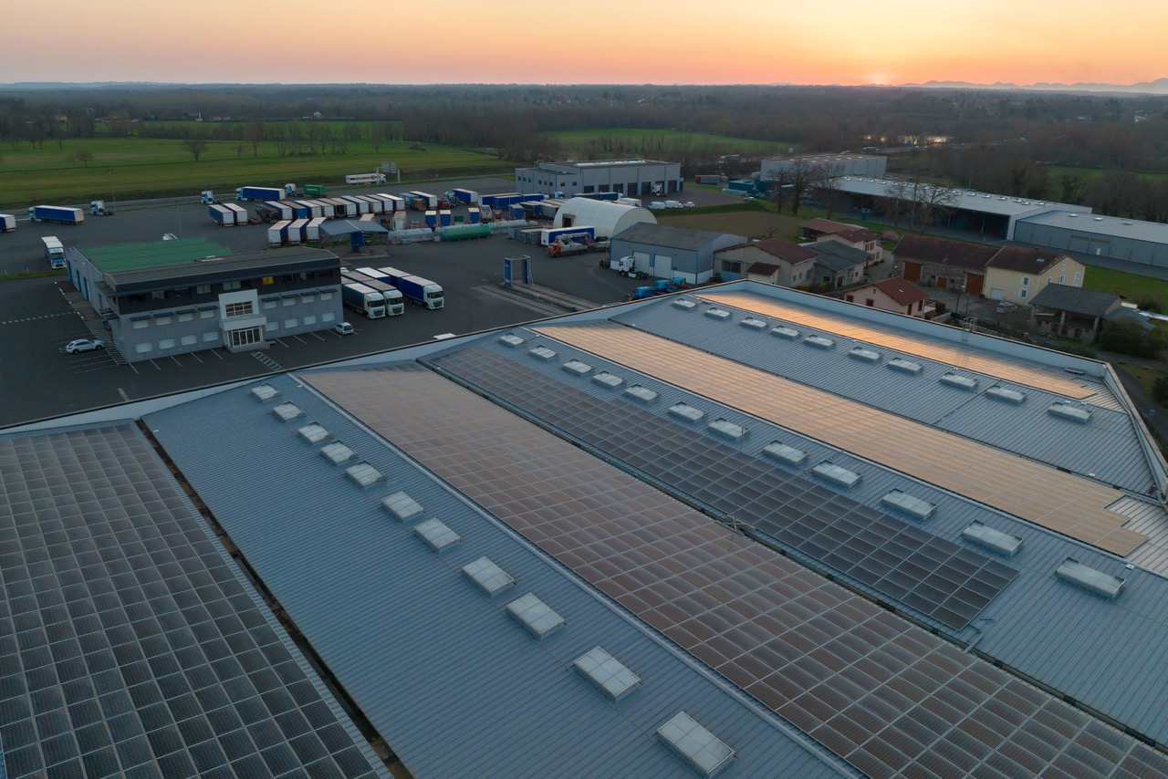 Aerial view of industrial buildings with solar panel roofs and trucks at sunset in a rural area.