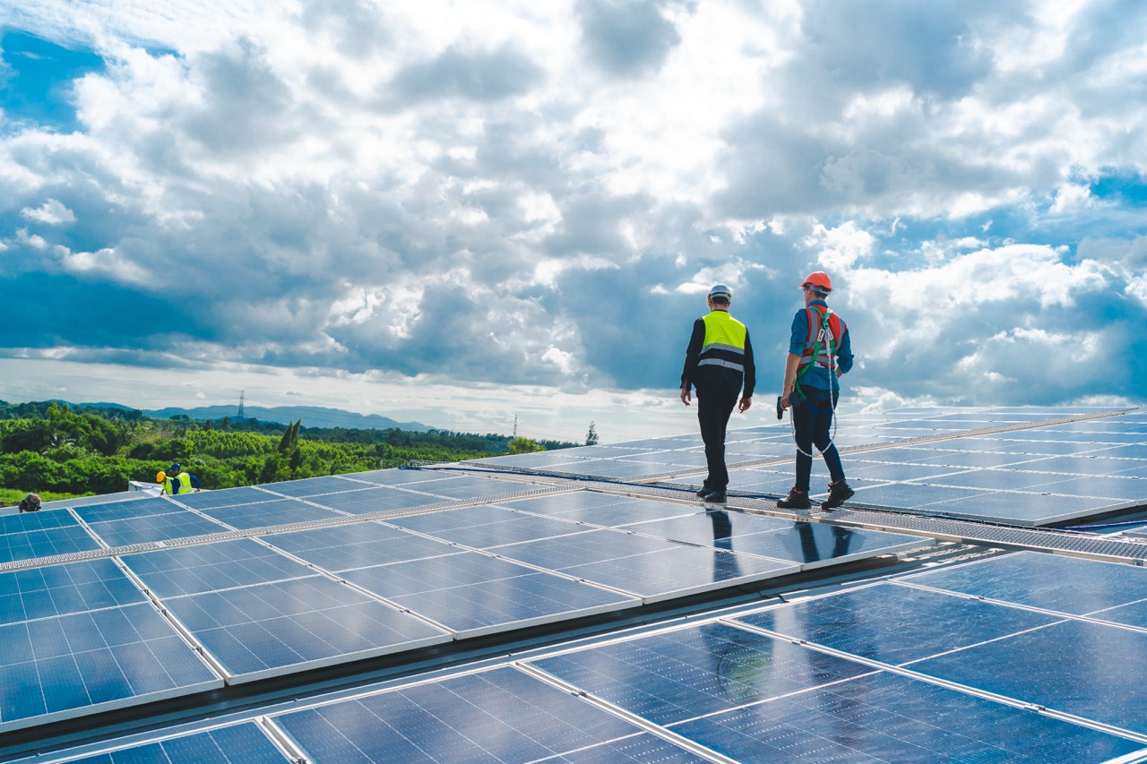 Two workers in safety gear inspecting solar panels under a cloudy sky with trees in the background.