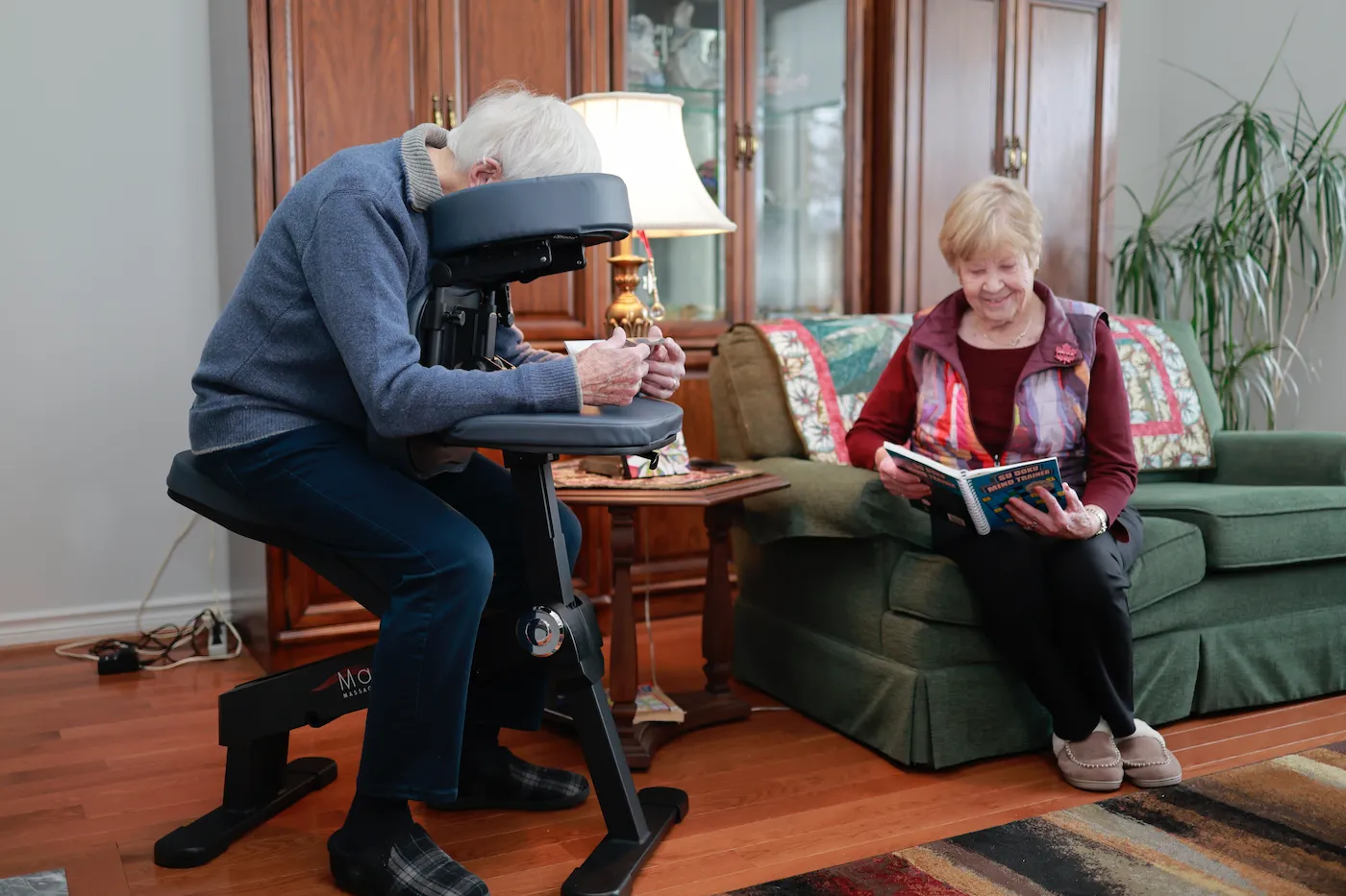 Elderly man receiving a seated massage while an elderly woman sits on a couch reading a book in a cozy living room.