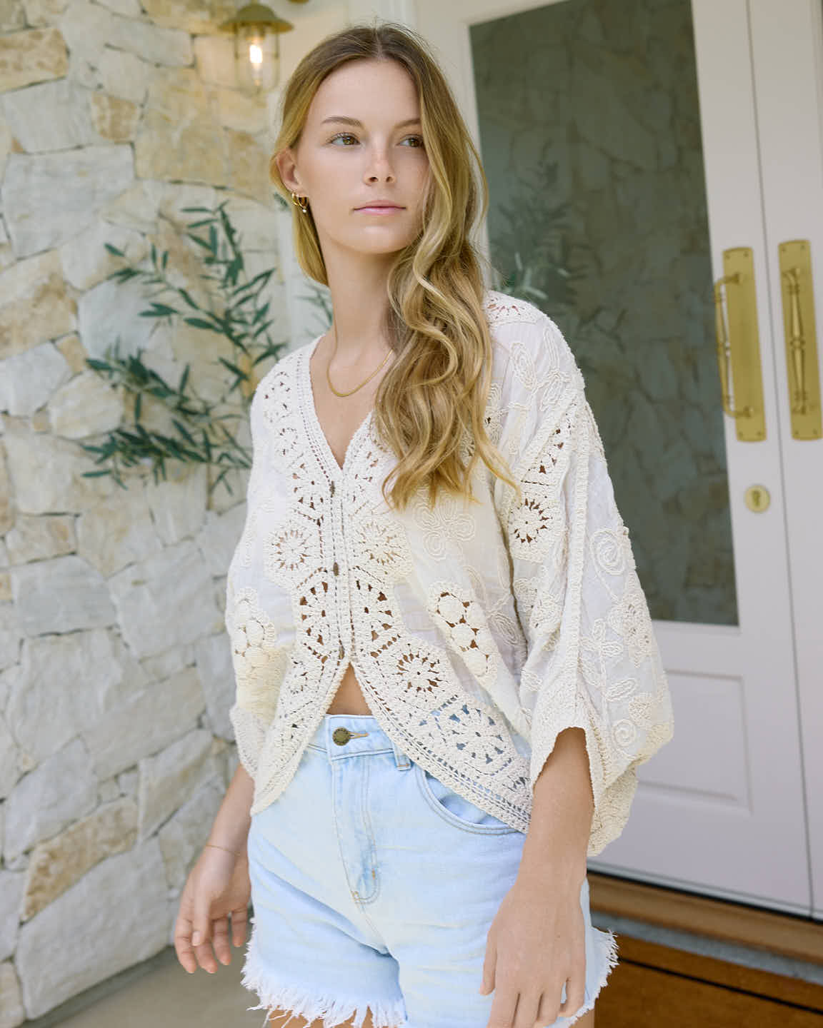 Young woman wearing a cream lace top and light blue denim shorts standing near a stone wall and glass door.