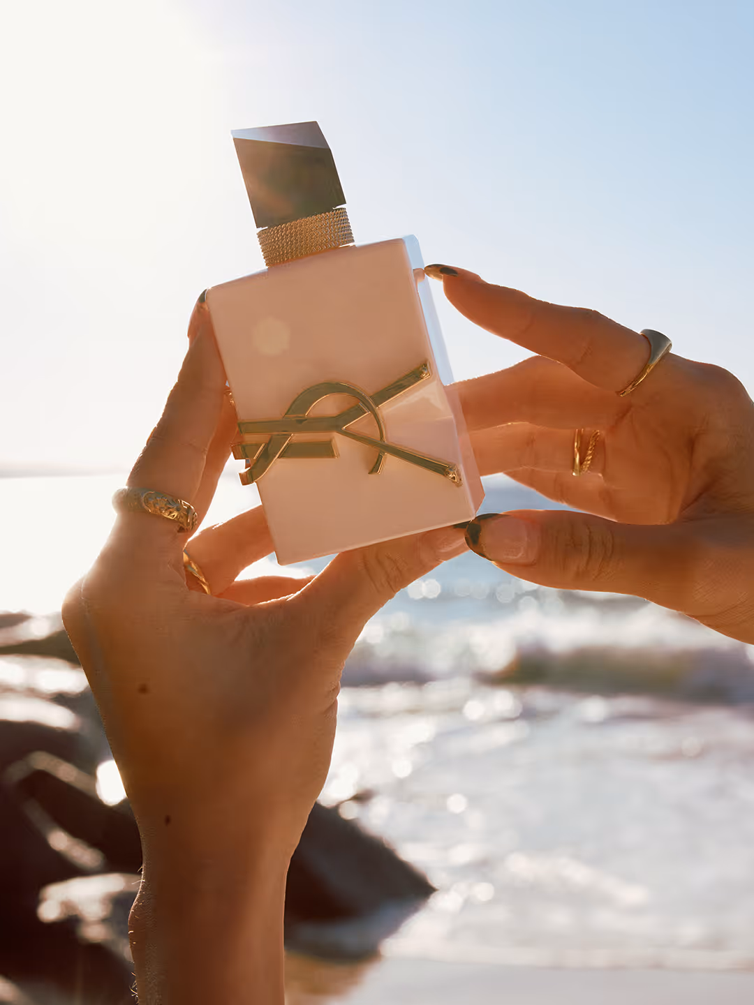Hands holding a beige YSL perfume bottle with a gold logo and black cap, with a sunlit beach background.