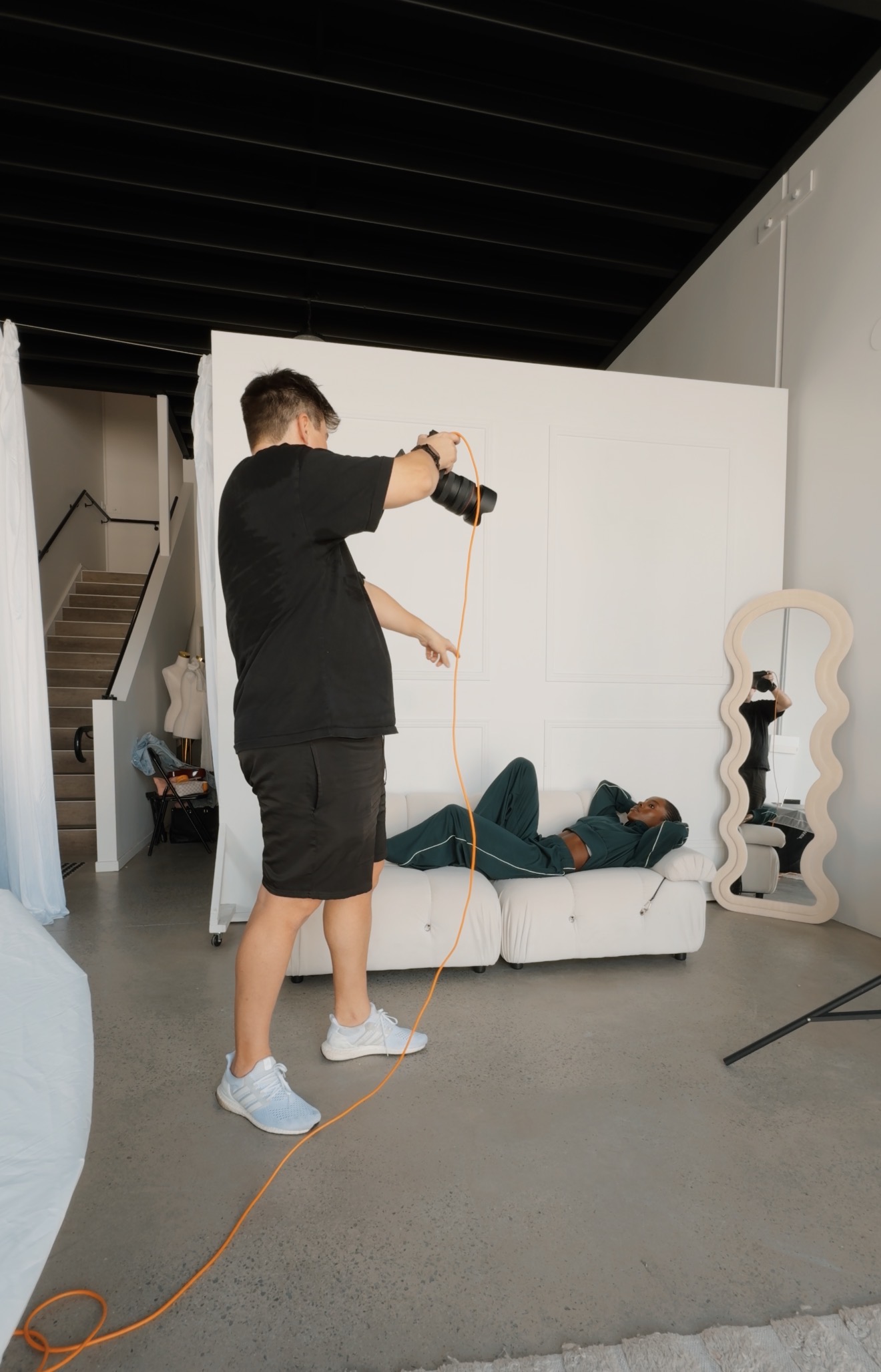 Photographer in black shirt and shorts taking photos of a woman lounging on a white couch in a studio.