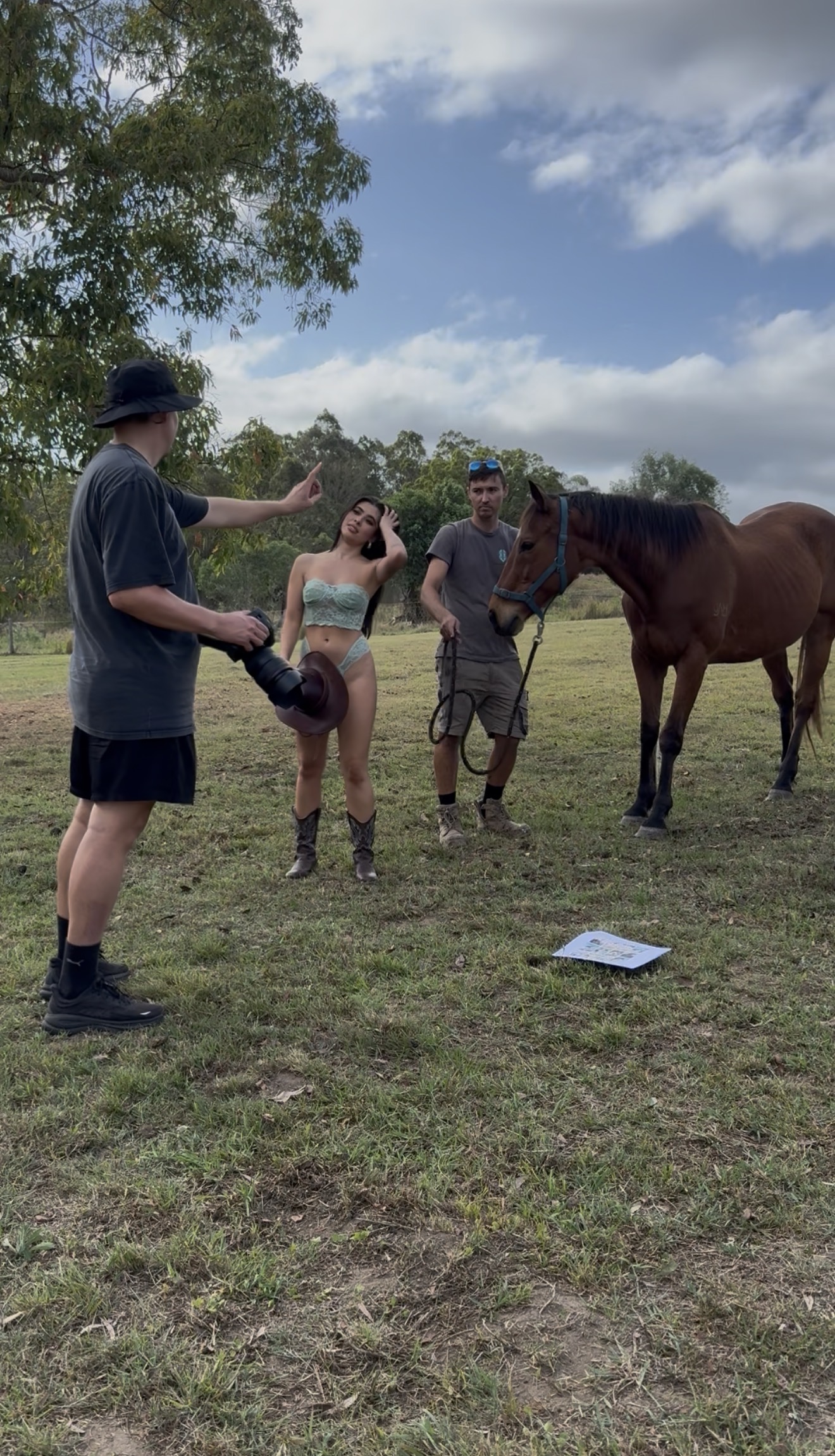 Photographer directing a woman in a green lingerie set and boots posing next to a man holding a brown horse on grassy field.