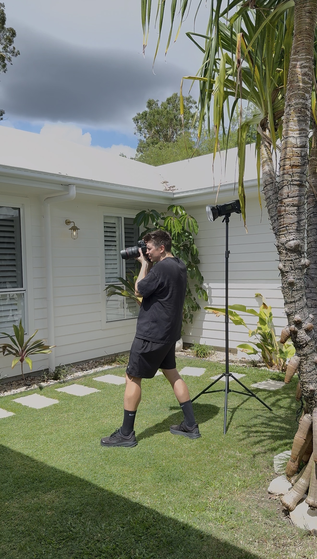 Person in black clothing taking a photo in a sunny backyard with tropical plants and a white house.