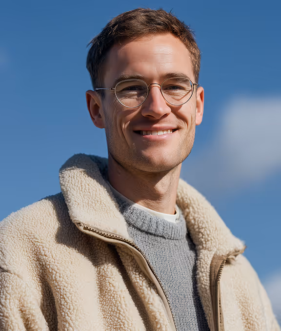 Smiling young man wearing round glasses, a beige sherpa jacket, and a gray sweater against a clear blue sky.