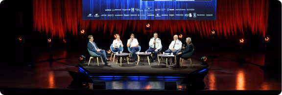 Panel de six personnes assises sur scène avec un grand écran en arrière-plan et un rideau rouge.
