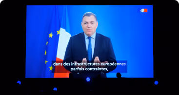Un homme en costume parlant devant les drapeaux européen et français sur un écran vidéo.