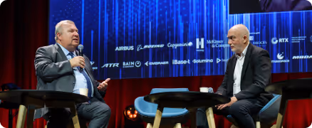 Deux hommes en costume discutent assis sur scène avec fond bleu et logos d'entreprises.