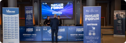 Un homme debout devant un comptoir d'inscription du Paris Air Forum avec écrans et panneaux bleus.