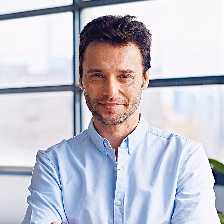 Confident man with dark hair and light stubble wearing a light blue button-up shirt standing with arms crossed in front of a window.